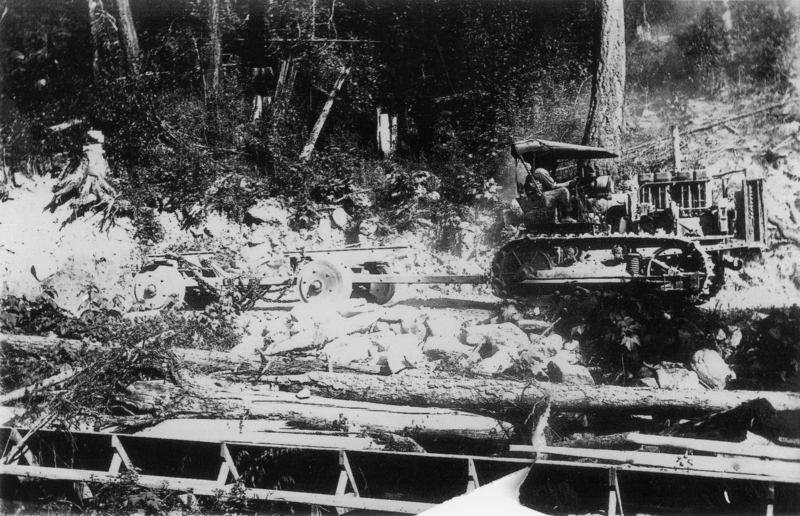 A person operating a tractor in a forested area, with fallen trees and rough terrain around them. The tractor appears to be pulling a piece of equipment on wheels through the rocky ground.