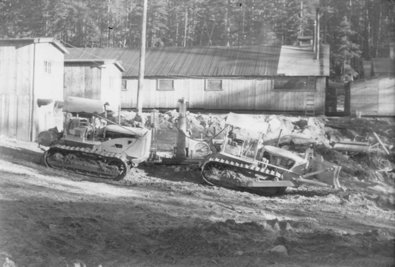 Two bulldozers working on a dirt path near a wooden building, surrounded by trees.