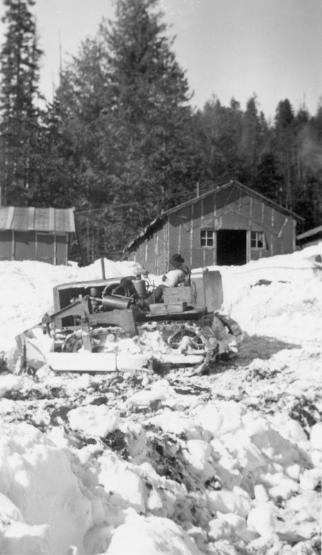 A person operating a bulldozer in a snowy area with two wooden buildings and tall trees in the background.