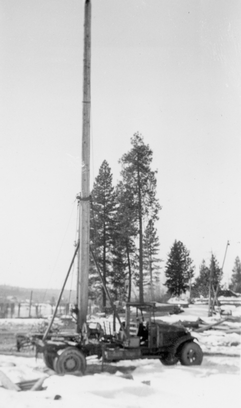 A truck with a tall, vertical mechanism attached, standing on a snowy landscape with trees and distant buildings in the background.