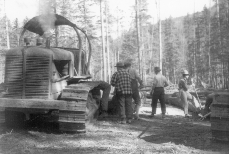 A group of men gathered around a bulldozer in a forest setting. Some men are wearing hats and are engaged in an activity involving logs or lumber. Trees and rough terrain are in the background.