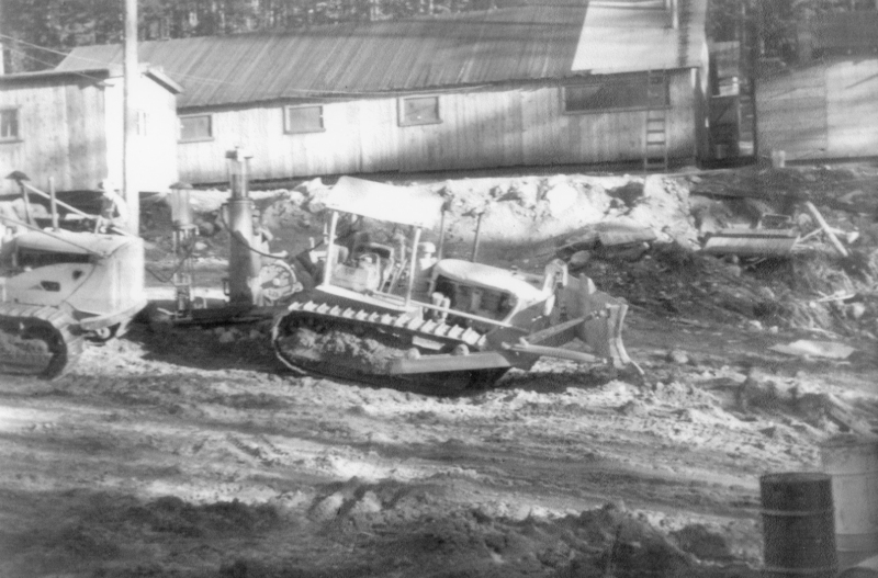 A construction scene with a bulldozer moving earth. There are two workers sitting in the bulldozer. A building is visible in the background, and a drum is located in the foreground.