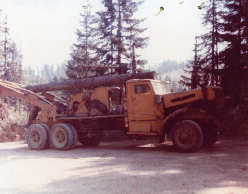 A large logging truck parked on a dirt road in a forested area, with tall trees visible in the background.
