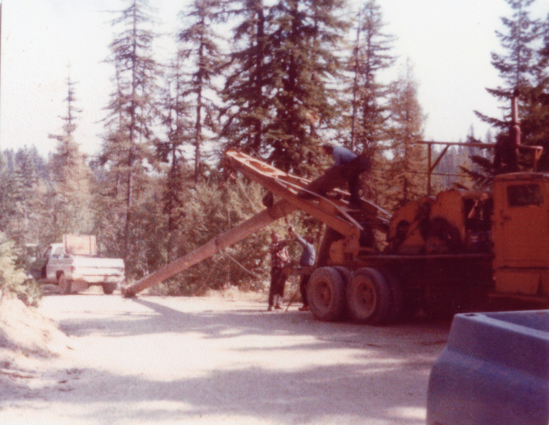 A person is climbing on a large piece of machinery with wheels, while another individual stands nearby on a dirt road surrounded by tall trees. A pickup truck is parked in the background.