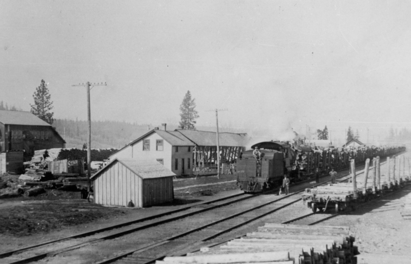 A train loaded with logs is on the tracks near a few buildings surrounded by stacks of lumber. A person is seen standing on the back of the train, and another person walks nearby. Tall trees and utility poles are visible in the background. The side of a structure near the center-left has text that reads: "STAR".