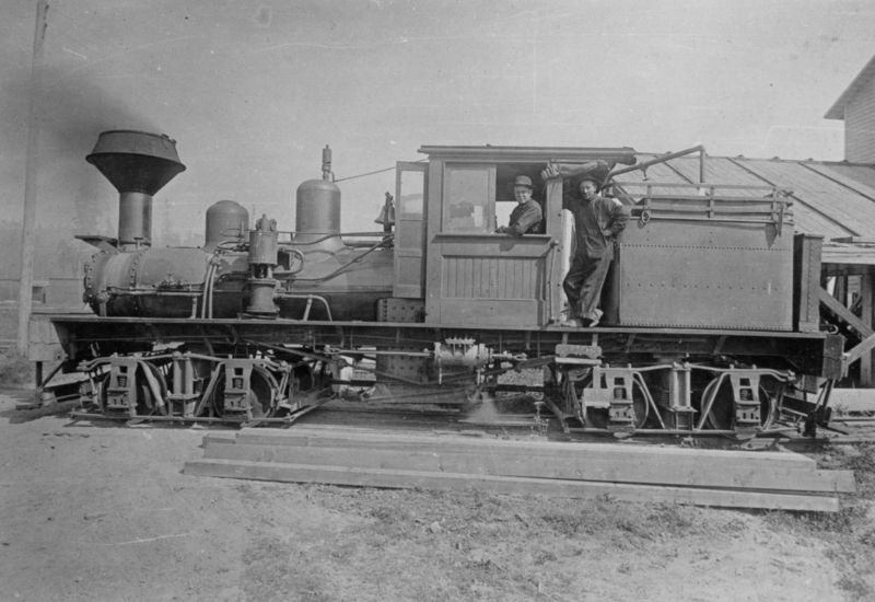A locomotive with two men in work attire, one standing in the cabin and the other leaning on the side, with a building in the background.