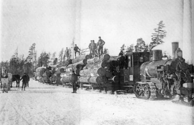 A steam locomotive is pulling a long load of large logs through a snowy landscape. Several men are standing on top of the logs and beside the train. There is a team of horses with a driver in the foreground. Tall trees can be seen in the background.