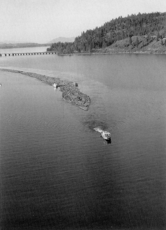 A river scene with a boat moving across the water. There is a large group of logs floating in the river, held together by a boom. A forested hillside borders the river, and a bridge spans the water in the distance.