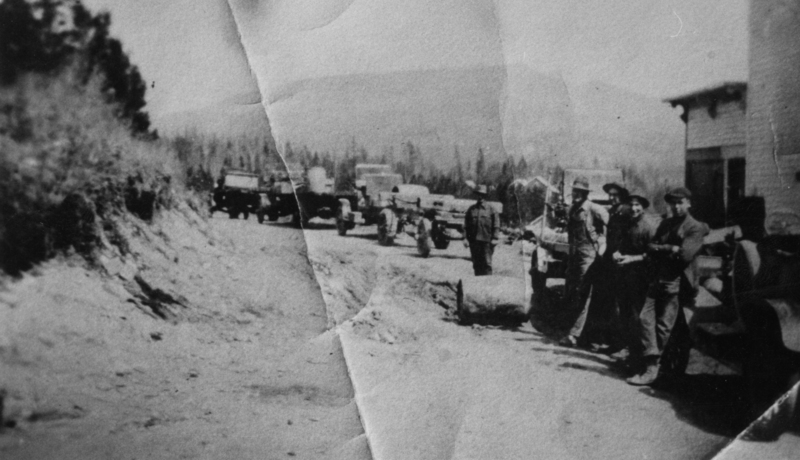 A group of men stands along a dirt road with several trucks carrying equipment or logs. Trees and distant hills are visible in the background, and there is a building on the right side.
