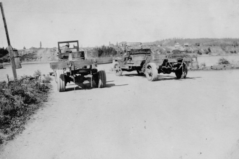 Two old trucks with open backs are parked on a dirt road. One truck has a seated driver. The area surrounding them is rural with fields, trees, and a few buildings visible in the background. A utility pole is on the left side near some vegetation.