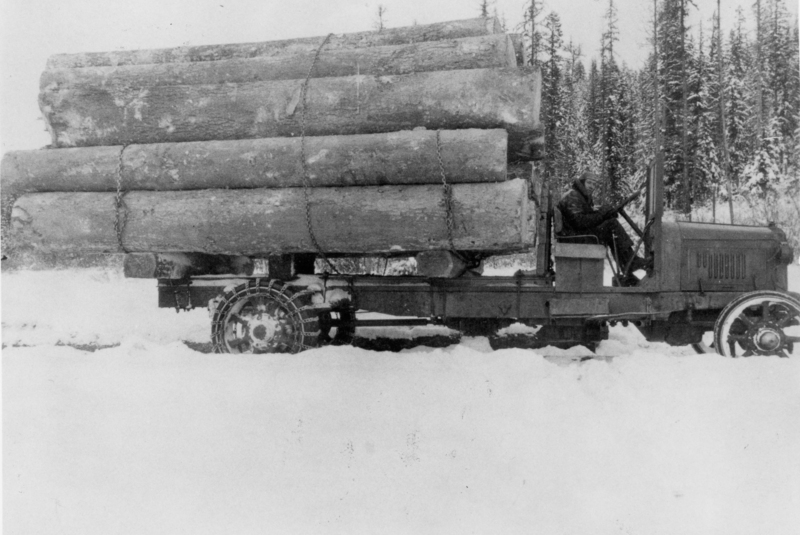 A tractor loaded with large logs, driven by a person, is moving through a snowy landscape with a forest in the background.