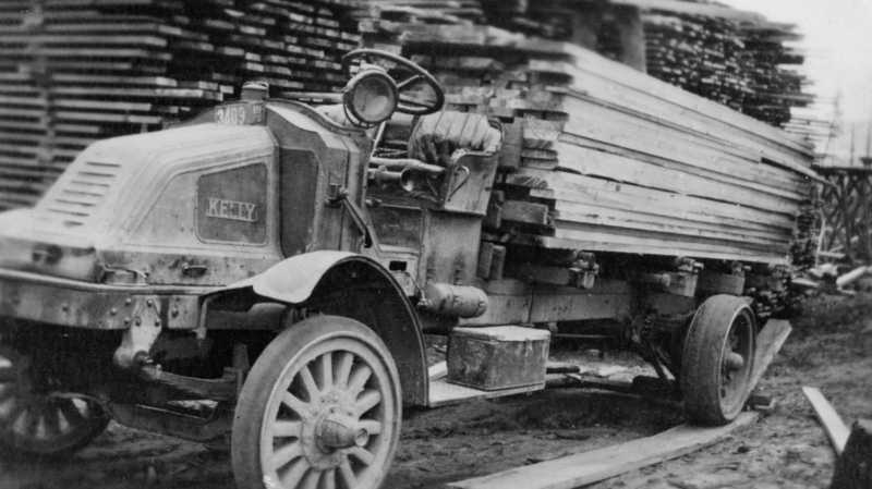 An old truck with wooden wheels carrying a large load of stacked wooden planks. The background is filled with more stacks of planks. The truck has one visible headlight and a steering wheel, and the text "Kelly" and "3409" are written on the vehicle.