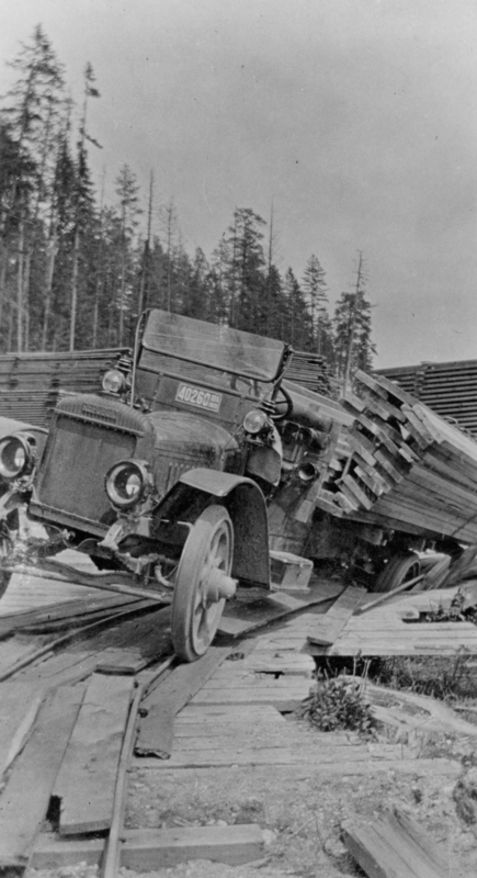 A truck carrying a large stack of lumber is tipped over, navigating a rough, uneven wooden path in a forested area. Tall trees are visible in the background. The license plate on the vehicle reads: "40260."