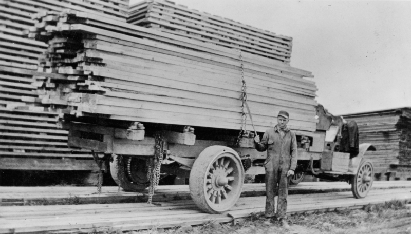A man standing beside a truck loaded with long stacks of lumber, surrounded by more piles of lumber. He holds a chain in one hand and is wearing a uniform and cap. The scene appears to be in a lumber yard.
