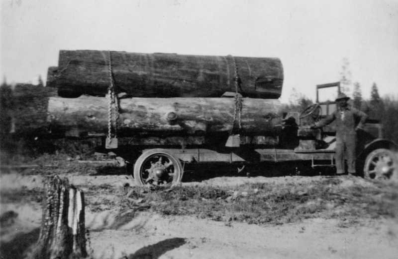 A man stands beside a truck loaded with large logs secured by chains. The scene is set in an outdoor, wooded area.