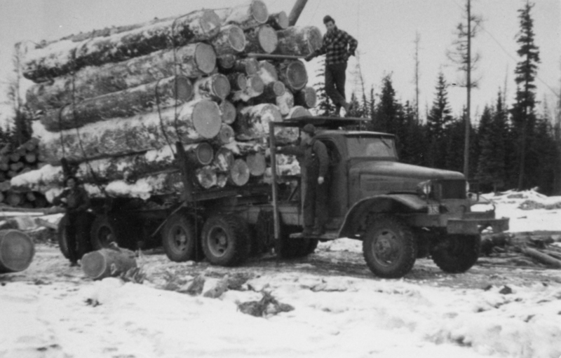 A truck loaded with large logs is parked in a snowy landscape. One person stands on top of the truck, securing the logs, while another person stands by the driver's door. Tall trees can be seen in the background.