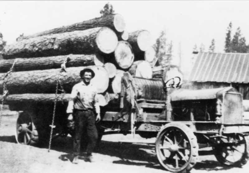 A man stands next to a truck loaded with large logs, wearing a shirt and pants. The background includes a building with a sloped roof and trees.