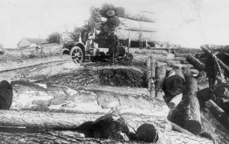A person driving a vintage truck loaded with large logs in a timber yard, surrounded by additional logs scattered around. A building is visible in the background.