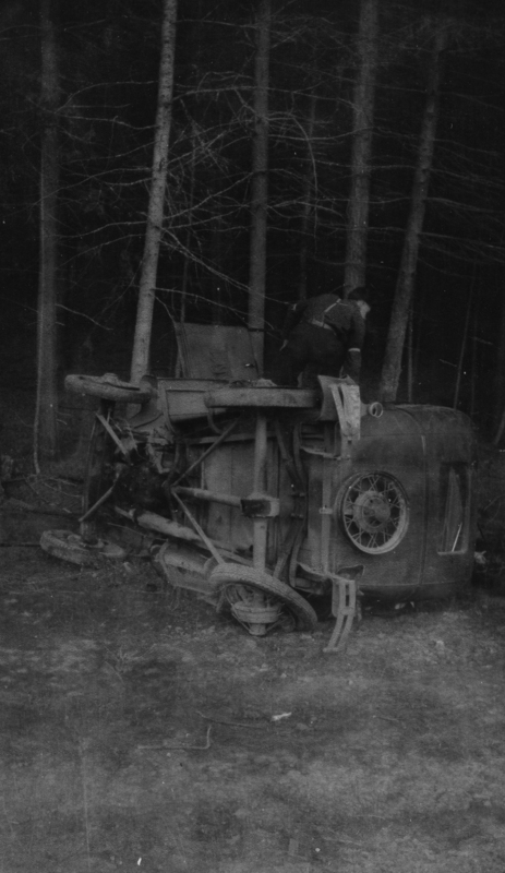 An overturned vehicle laying on its side in a forested area, with a person in uniform inspecting or reaching inside it.