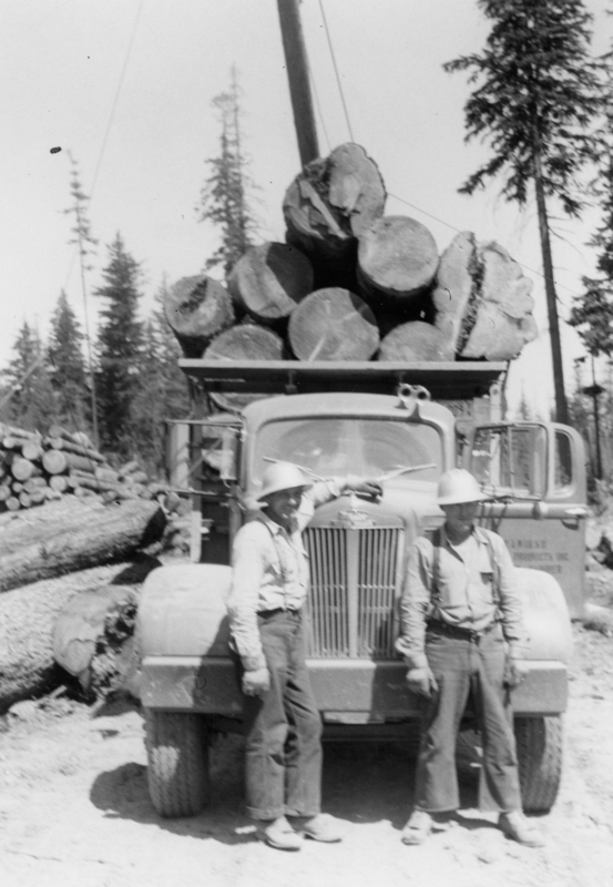 Two men wearing hard hats standing in front of a truck loaded with large logs. Trees are visible in the background.