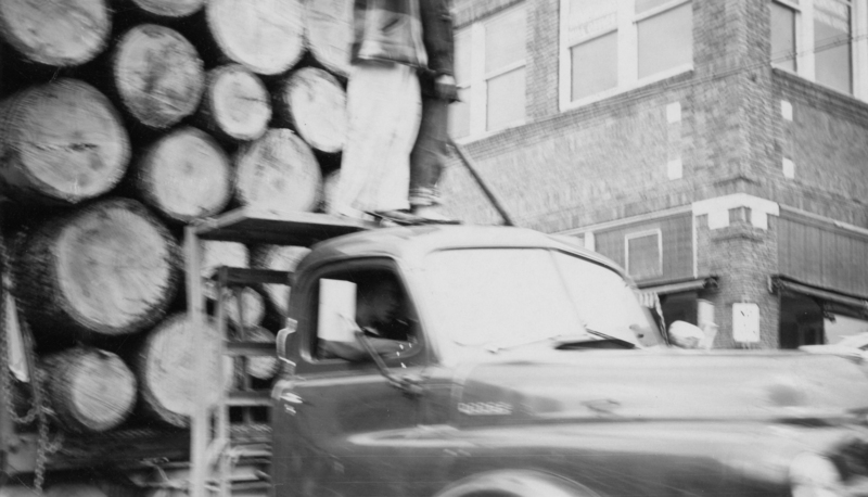 A truck loaded with large logs is parked near a brick building. A person is sitting inside the truck cabin, while another person stands on the truck platform near the logs.