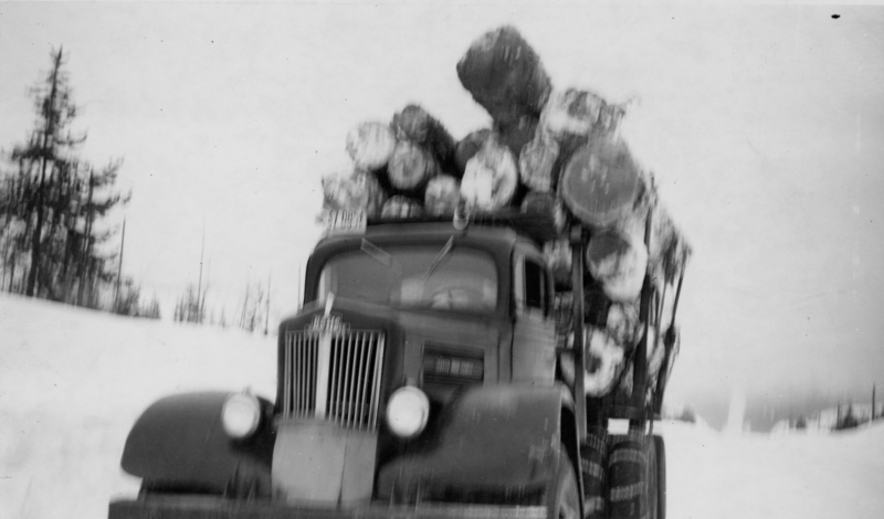 A truck loaded with large logs, traveling down a snowy road, with trees visible in the background.