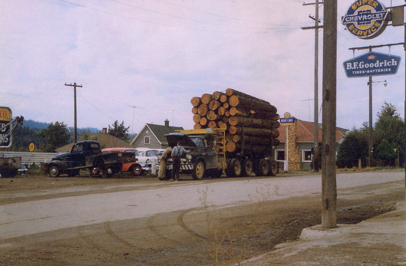 A large truck loaded with logs parked on a roadside, with two people inspecting the front of the truck. Nearby are two vintage cars parked beside each other. Sign near top right reads "Chevron SUPER SERVICE".  Below that, another sign reads "B.F. Goodrich TIRES • BATTERIES". A sign in the background near the center of the image reads "USED CARS".