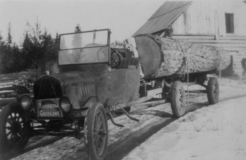 An old truck with a sign reading "Rainbow Gasoline" is parked in a snowy area next to a wooden building. It is pulling a large log secured to its trailer. Trees are visible in the background.