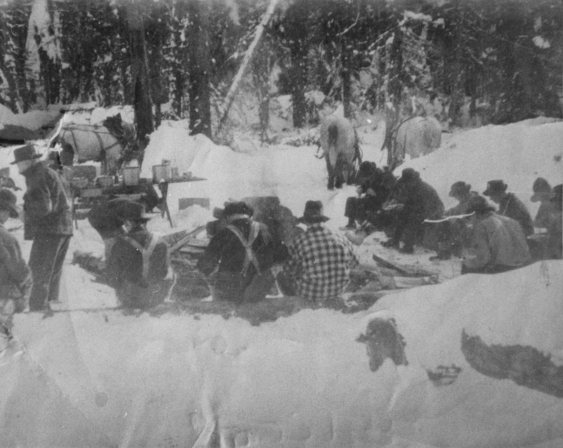 A group of people wearing hats sitting and standing in a snowy forest clearing, with two horses partly visible in the background and a table with pots and equipment nearby. Some people are huddled close to what appears to be a fire.