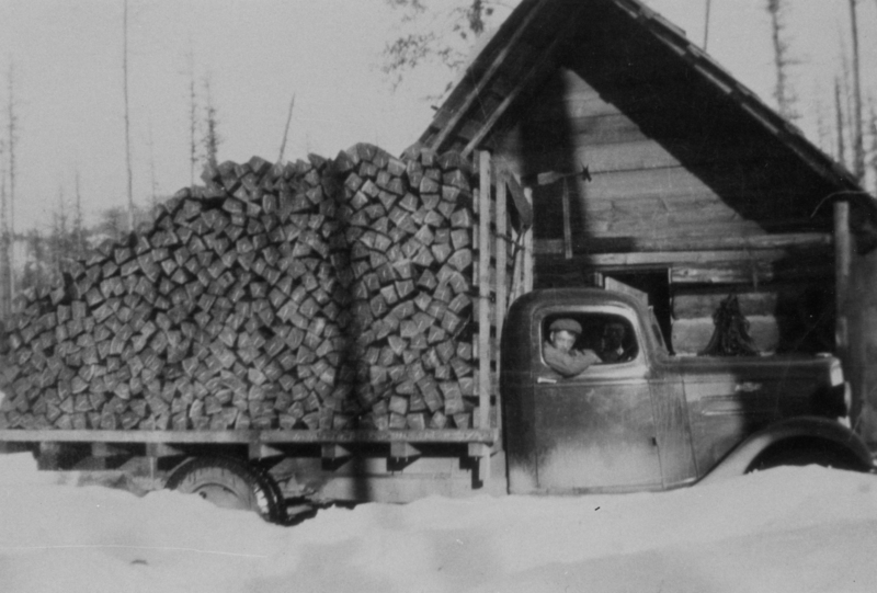 A person sitting in the cab of a truck loaded with stacked firewood, parked next to a wooden building in a snowy area.