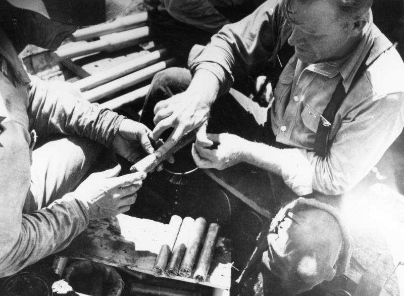 Two men examining a stick of dynamite laid out on a wooden surface. One man wears a buttoned shirt with suspenders.