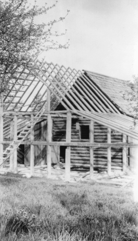 A partially constructed wooden building with visible framing and a partially completed roof, surrounded by trees and grass. A ladder leans against the structure.