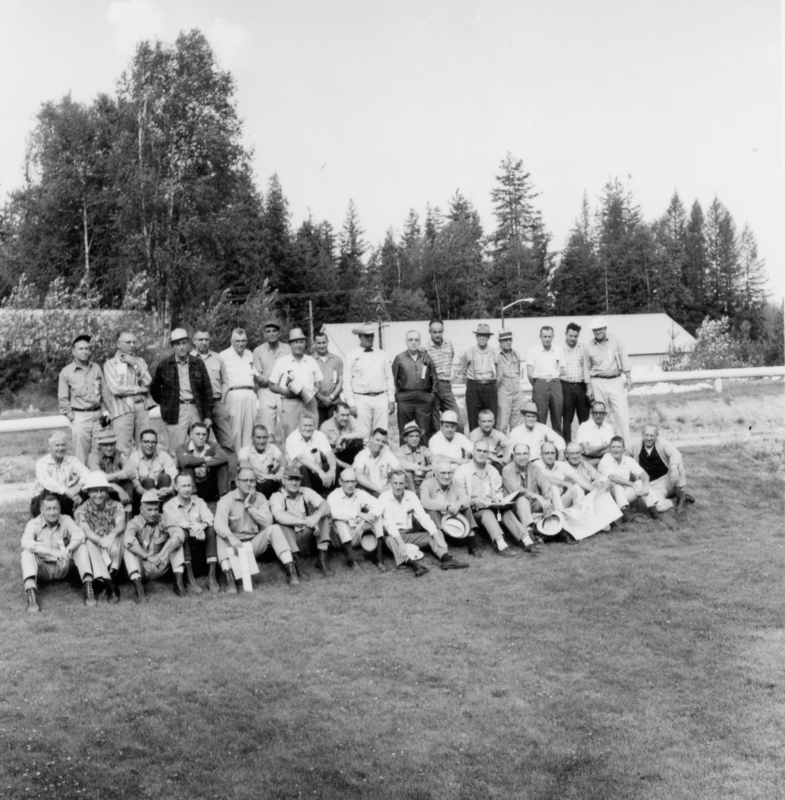 A large group of men, some wearing military uniforms, posing together in rows on a grassy field with trees in the background.