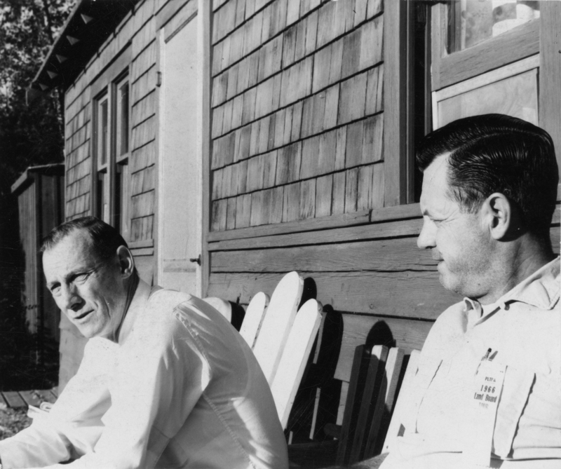 Two men are sitting outside next to a wooden building with shingle siding. One man is wearing a collared shirt and appears to be speaking, while the other, also in a collared shirt, is looking at him. They are seated in wooden chairs. One wears a pendant which reads: "PLTPA (Priest Lake Timber Protection Association), 1966 Land Board Tour".