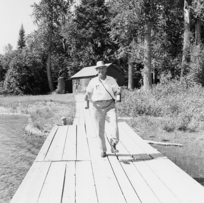 A man wearing a hat and casual clothing walks along a wooden dock carrying two bottles. There are trees and a small cabin in the background.