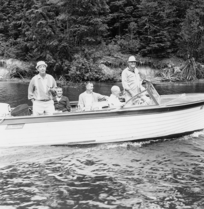 A group of five people on a boat. Two men are standing, and three are seated. Water and a forested shore are visible in the background. A label near the bow of the boat reads "Thompson."