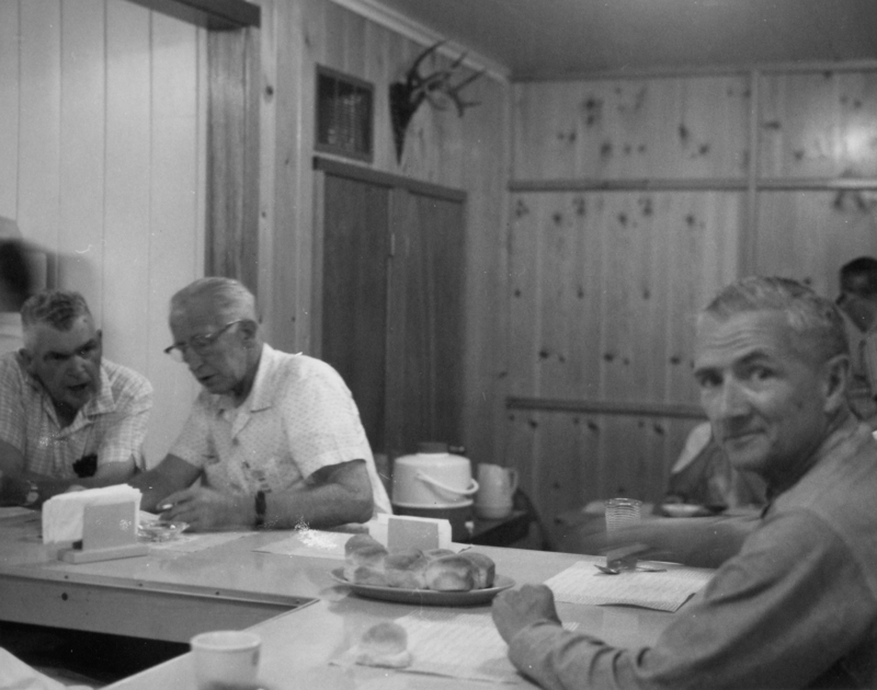 Three men sit at a table in a room with wooden walls. Two of them are engaged in conversation, while the third faces the camera. A table holds a plate of rolls, cups, and some paper items. A mounted deer head is visible on the wall.