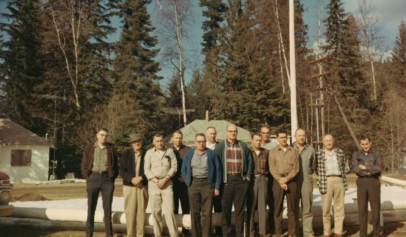 A group of twelve men standing together outdoors in front of a forest. Some are wearing jackets and hats, and a house with a green roof is visible in the background.