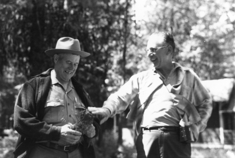 Two men are standing outdoors, smiling. One man is wearing a hat and holding a small bundle, while the other man, dressed casually, is reaching toward it. Trees are visible in the background.