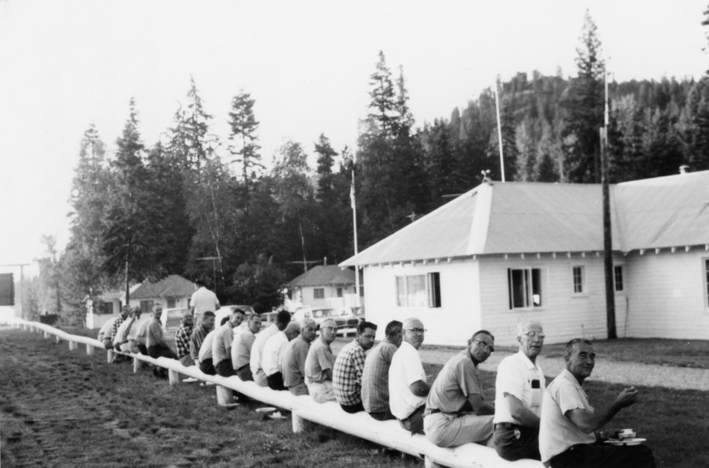 A group of men sitting on a long bench facing away from the camera, with a few looking back. They are outside near a building with a sloped roof, surrounded by trees. Some men hold plates, having a meal.