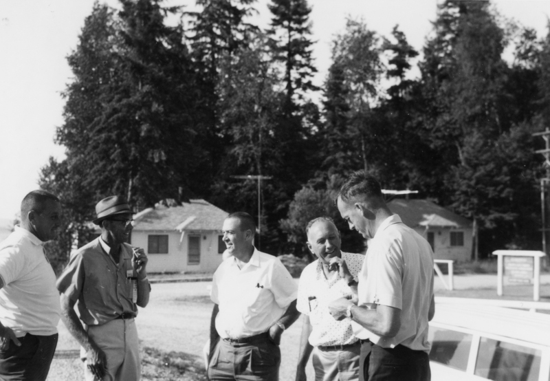 Five men standing outdoors near trees and houses, engaged in conversation. One man is holding a notepad and pen, while another has a cigarette. They appear to be in a casual setting.