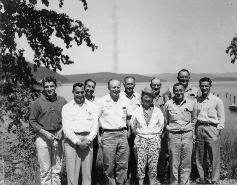 A group of nine people, consisting of eight men and one woman, standing outdoors near a body of water. Some men are in casual shirts or uniforms. The woman is wearing a hat and patterned pants. Trees and hills are visible in the background.