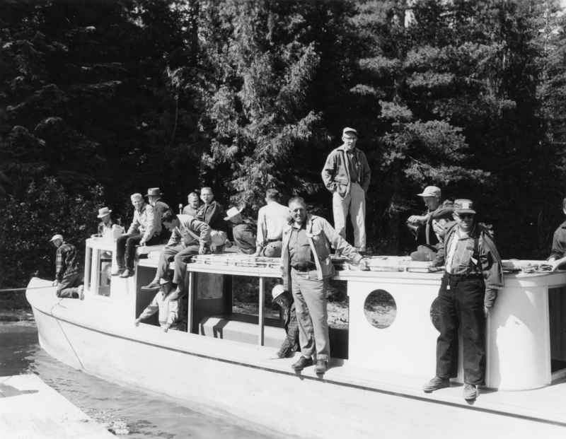 A group of men sitting and standing on a boat surrounded by trees. Some are seated on the roof while others stand on the deck, appearing relaxed and engaged with each other.