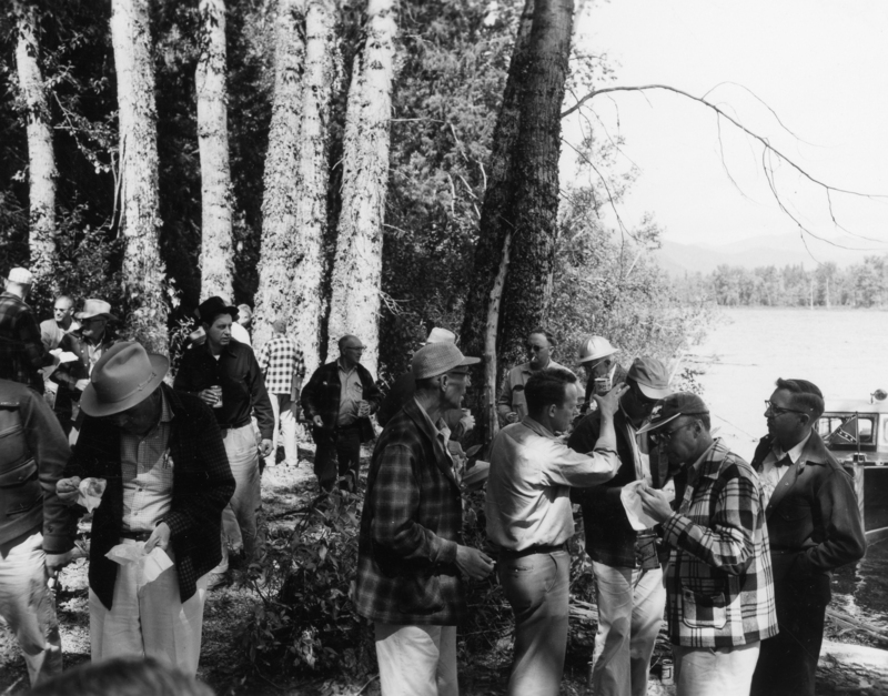 A group of men gathered near a lakeside surrounded by trees. Some are wearing hats and holding food or drinks. A few stand near a vehicle by the water. The scene appears relaxed, with people talking and socializing.