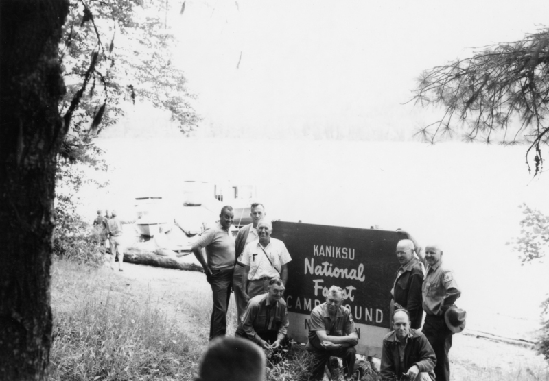 A group of men is gathered around a sign that reads "Kaniksu National Forest Campground." They are standing and kneeling near a lake with trees surrounding the area. In the background, a few people stand near some boats by the water.