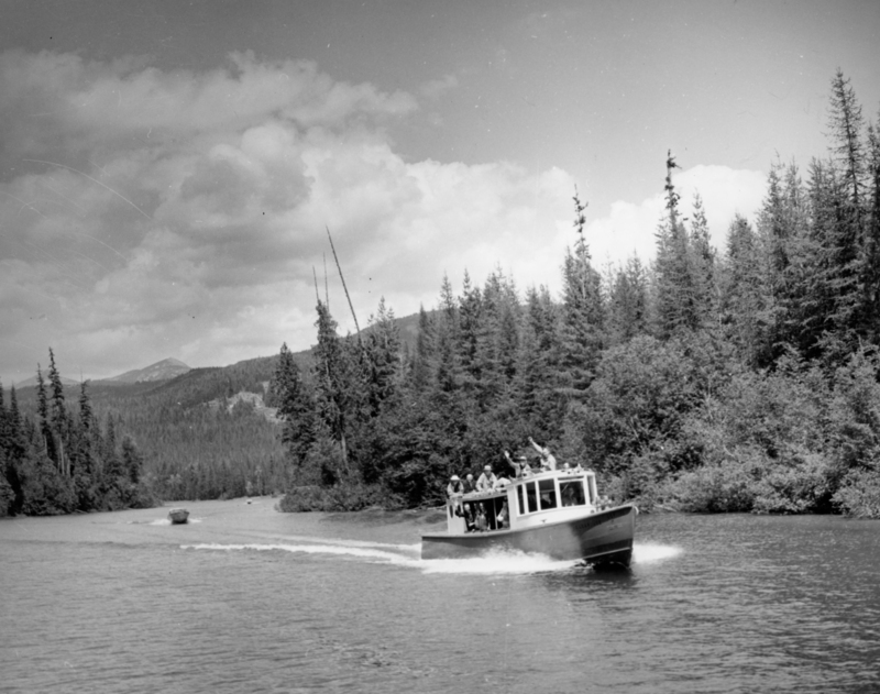A group of people on a boat traveling through a river surrounded by dense forest, with mountains visible in the distance and a cloudy sky overhead.