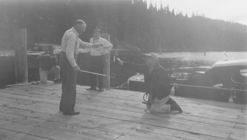 A man kneeling on a wooden dock extends a bottle towards another man standing and holding a tool resembling a hook. Two other individuals stand behind them with one leaning on a post, and trees and water are visible in the background.