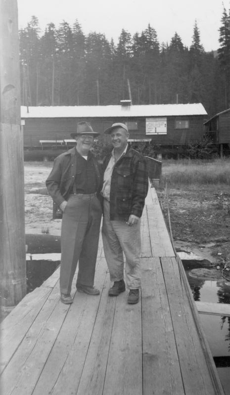 Two men standing arm in arm on a wooden dock. Both are wearing hats and jackets. A sign in the background reads: "THE DIAMOND MATCH CO. SAFETY RECORD, DAYS SINCE LAST INJURY, INJURIES THIS YEAR, JAMMERS, TRUCKS, SAWS".