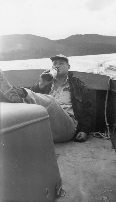 A man sitting in a boat with his legs stretched out, drinking from a bottle. He is wearing a cap and casual clothing, with a body of water and hills visible in the background.
