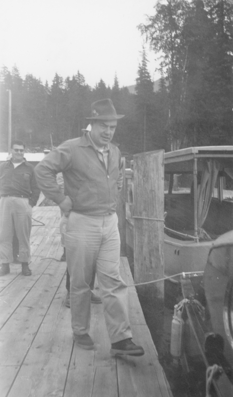 A man wearing a hat stands on a wooden dock next to a small boat. Another man is seen in the background wearing sunglasses. Tall trees are visible behind them.